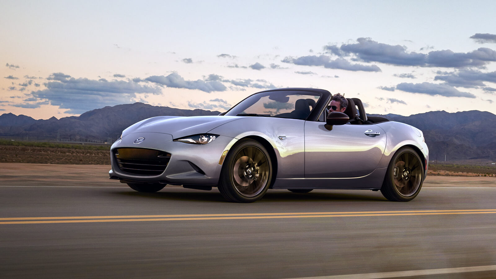 Une Mazda MX-5 ST de couleur gris mécanique métallisé avec le toit souple ouvert s’éloigne sur l’autoroute au-delà d’une chaîne de montagnes sous un ciel bleu et la lumière du soir.