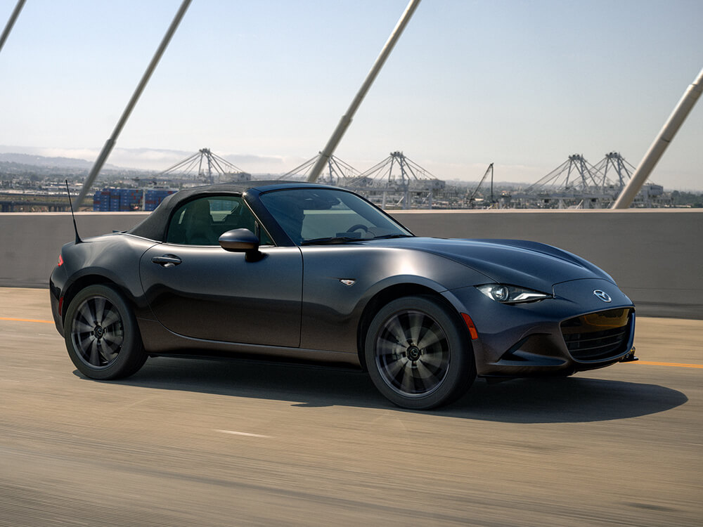 Une MX-5 ST gris mécanique métallisé avec le toit fermé traverse une passerelle avec un quartier industriel en arrière-plan.