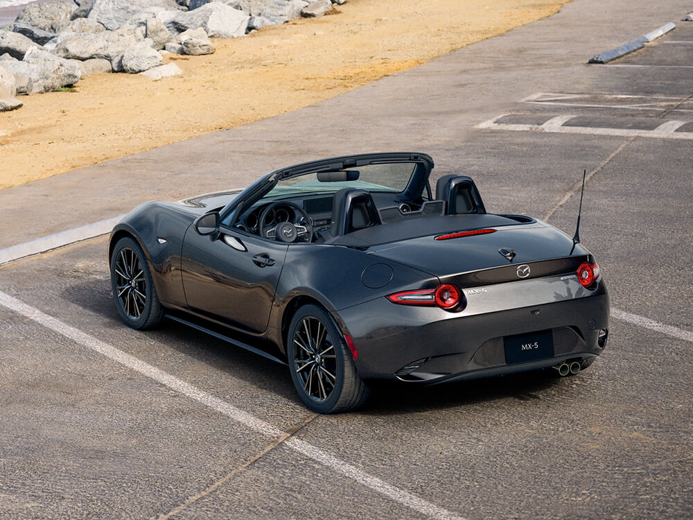 Vue arrière en plongée d’une MX-5 ST gris mécanique métallisé avec le toit baissé, garée dans un stationnement en bord de mer.