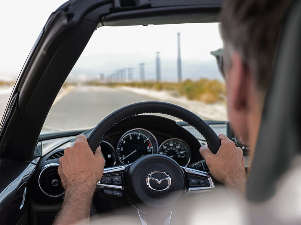 Vue arrière du conducteur d’une MX-5 ST; les mains sur le volant, il regarde une route dégagée bordée de poteaux électriques.