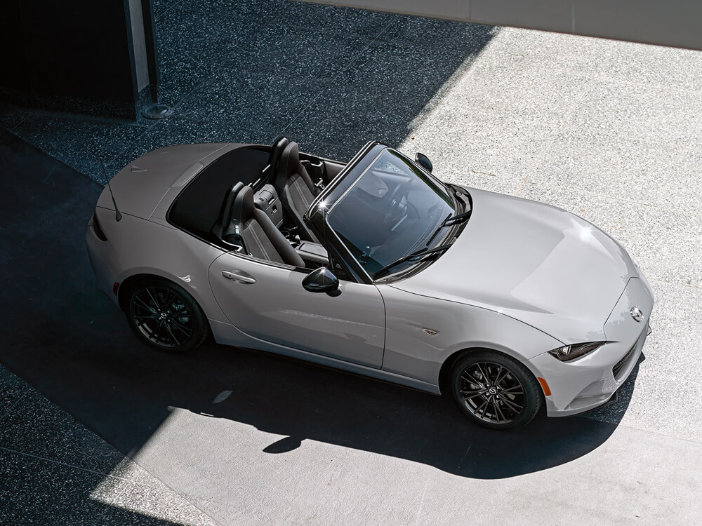 Vue côté passager d’une MX-5 ST gris aéro métallisé garée avec le toit baissé; l’arrière est assombri par l’ombre d’un bâtiment environnant tandis que l’avant brille sous le soleil de midi.