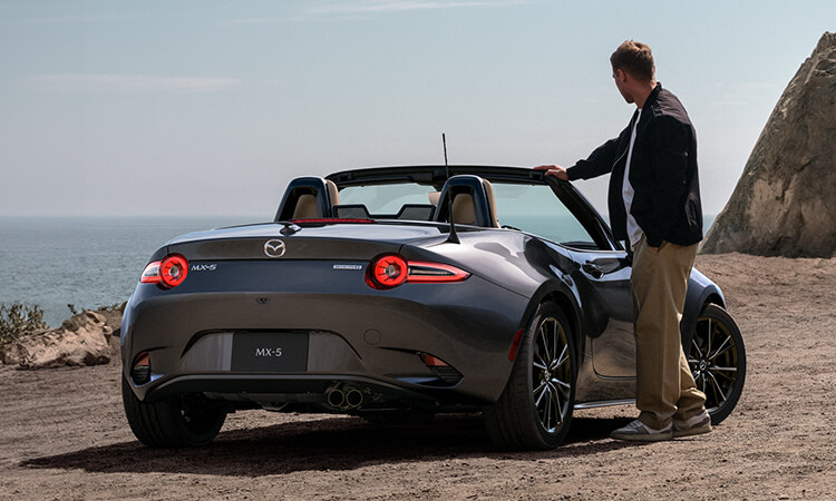 Vue de l’arrière d’une MX-5 gris mécanique métallisé avec le toit baissé; le conducteur est adossé au pare-brise et regarde un grand lac.