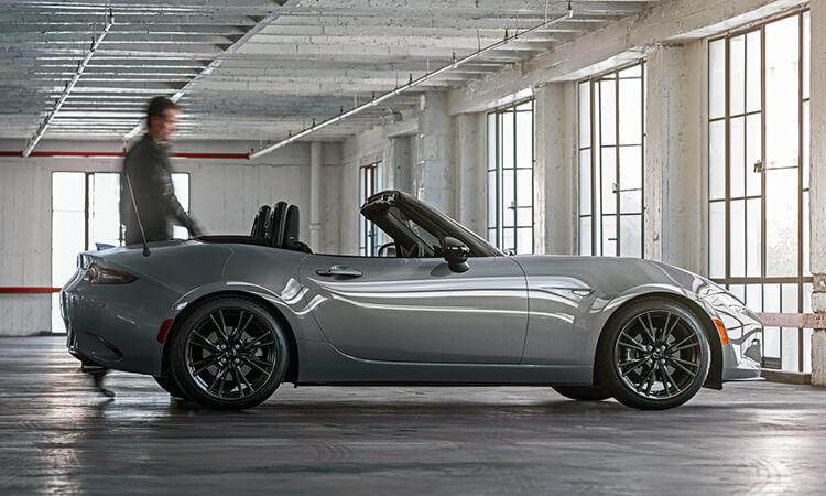 Passenger’s side profile of an MX-5 parked in a well sunlit parking structure, a driver swiftly approaches from behind running his hand along the edge of the car.