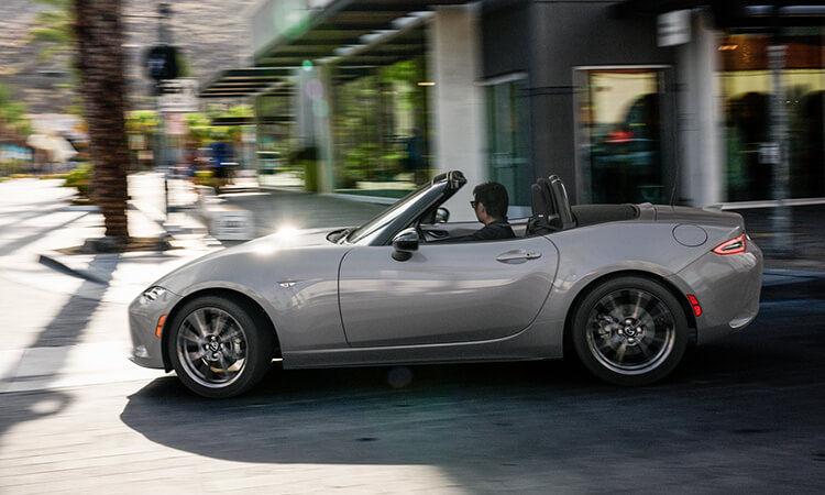 Vue de profil du conducteur d’une MX-5 gris mécanique métallisé qui tourne à droite dans une rue urbaine. 