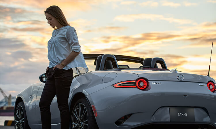 Une femme à côté d’une MX-5 gris aéro métallisé au coucher du soleil.