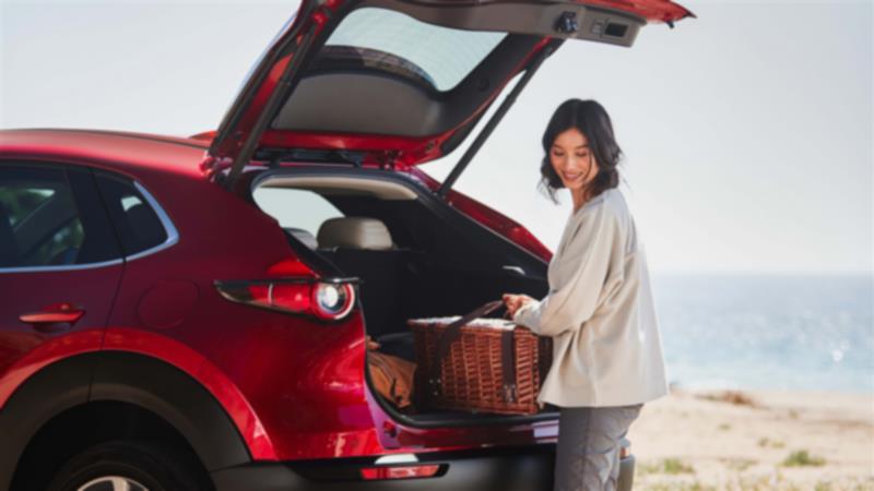 Un CX-30 rouge vibrant cristal métallisé garé au bord d’un lac. Une femme retire un panier de pique-nique/plage en osier par le hayon ouvert.