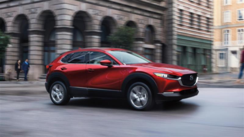 Gros plan de profil 7/8 d’un Mazda CX-30 rouge vibrant cristal métallisé traversant la ville, avec de majestueux bâtiments en pierre en arrière-plan. 