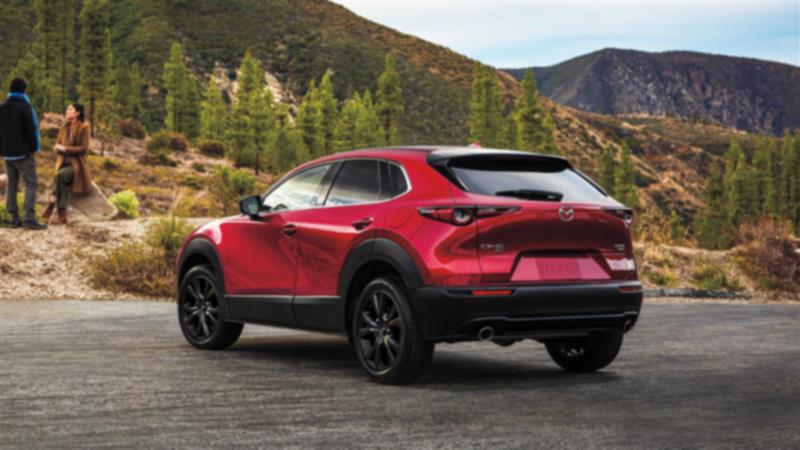 Plan de ¾ arrière d’un CX-30 rouge vibrant cristal métallisé garé au bord d’une route avec des montagnes et des arbres en arrière-plan. 