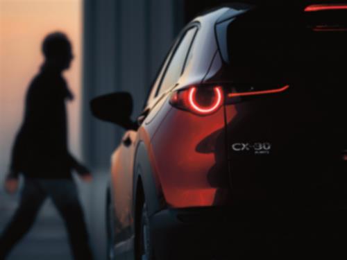 Close up of rear driver’s side of Soul Red Crystal Metallic CX-30 parked at dusk, focusing on CX-30 and AWD badge. Red tailight is illuminated. In the background, an out of focus silhouette of a man walking.