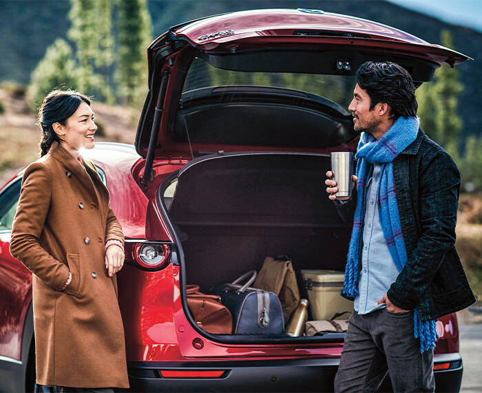 A couple converses behind their CX-30 with rear liftgate open. Dufflebags, cooler, and other items fill the cargo space.