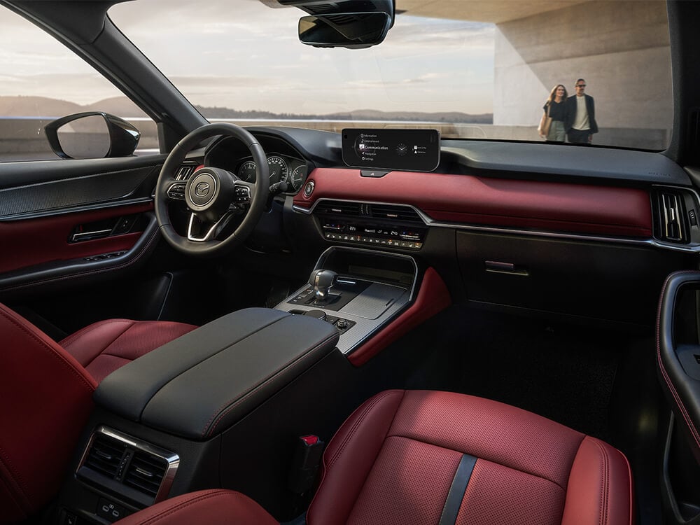 Interior shot looking out the windshield from the passenger’s seat inside a Garnet Red Leather interior CX-70, a couple walks back to their vehicle.