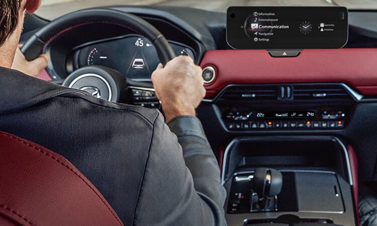 Vue par-dessus l’épaule du conducteur d’un CX-70 montrant le tableau de bord et le siège en cuir rouge grenat.