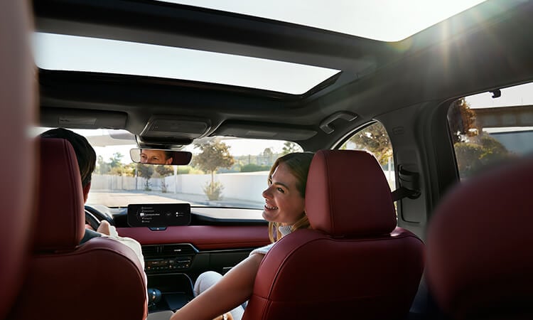 Shot from behind the second row, Garnet Red Leather interior of the CX-70. The passenger smiles wide at the driver.