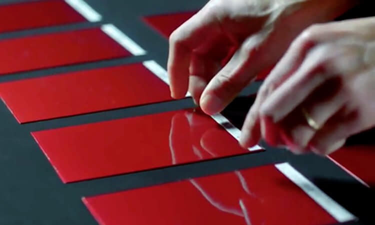 Close up of hands carefully grasping a red paint swatch on a black table under harsh light among other similarly coloured swatches.