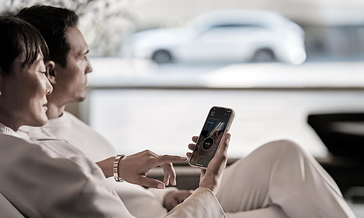 A couple sits together in their home, one on her phone with the MyMazda app open. A Rhodium White Metallic CX-70 parked in the background. 