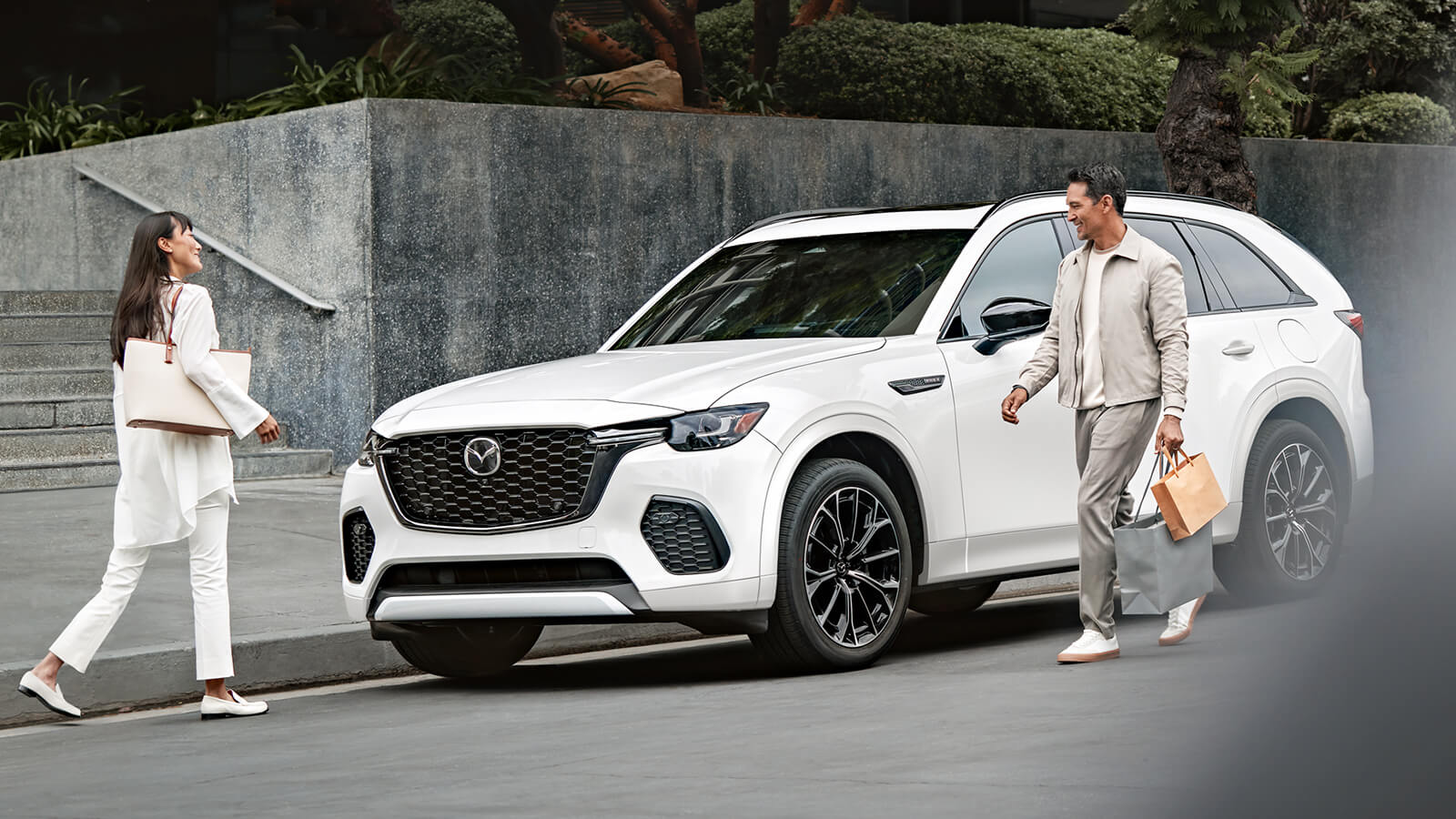 Man steps out of Rhodium White Metallic CX-70 with shopping bags and walks toward smiling woman.