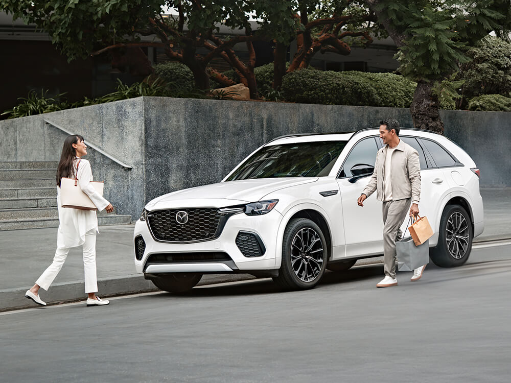 Man steps out of Rhodium White Metallic CX-70 with shopping bags and walks toward smiling woman.