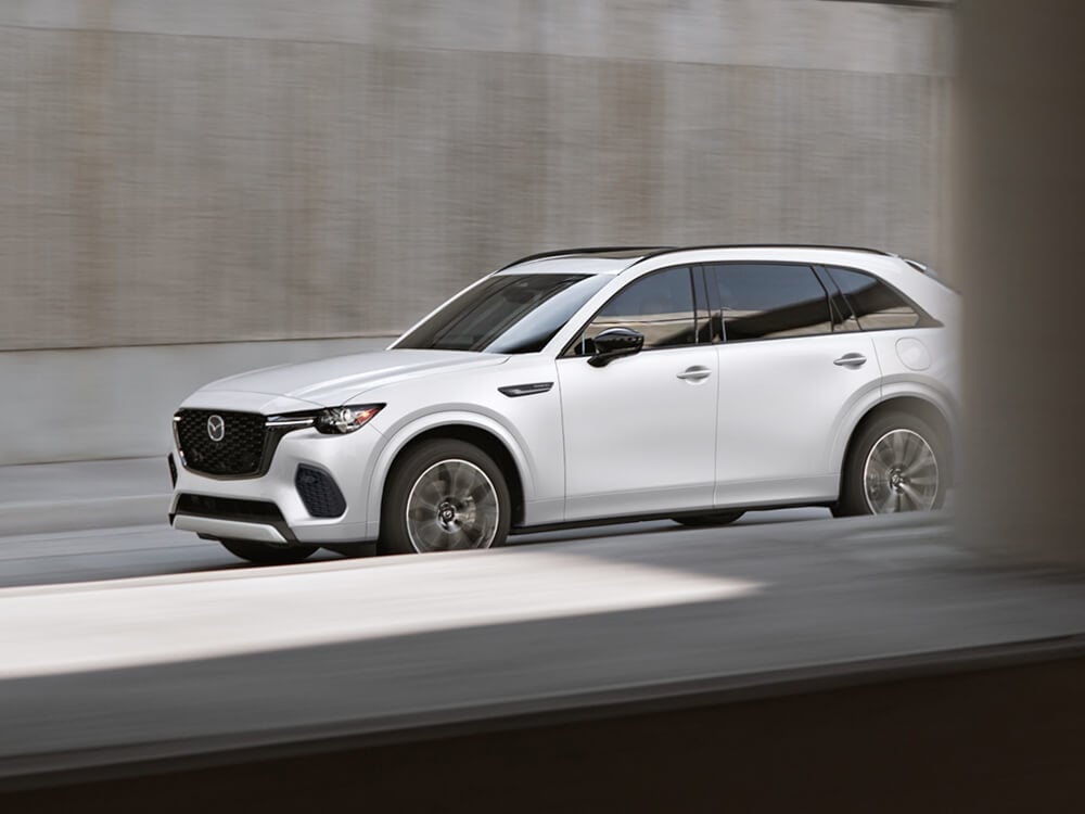 Driver’s side shot of a Rhodium White Metallic CX-70 driving in the shade of a concrete overpass.