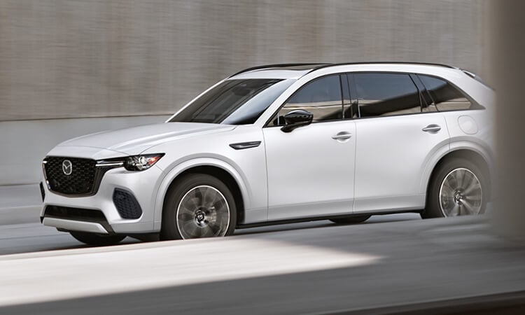 Driver’s side shot of a Rhodium White Metallic CX-70 driving in the shade of a concrete overpass. 